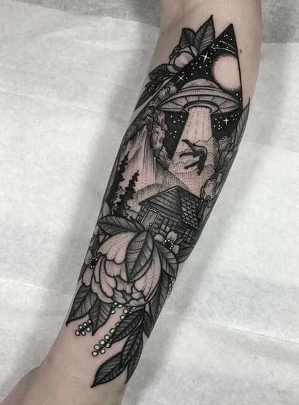 Tatuagem blackwork no antebraço com cena de abdução alienígena sobre uma cabana na floresta, emoldurada por flores e folhas em estilo dotwork.