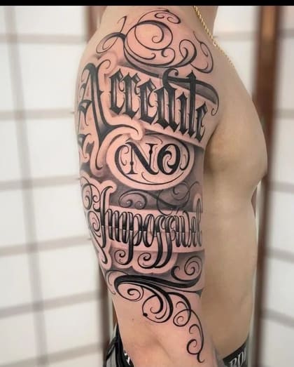 Tatuagem lettering 'Acredite no Impossível' em estilo chicano blackwork no braço de um homem, com sombreamento e ornamentos.