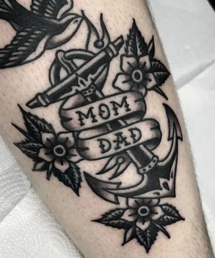 Tatuagem Old School de âncora com banner 'Mom Dad', flores e andorinha em preto e cinza, simbolizando amor familiar e lealdade.
