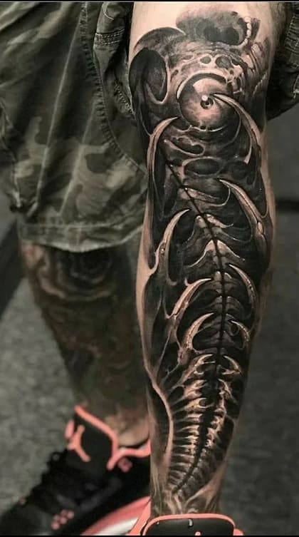Tatuagem bio-orgânica detalhada na perna, em preto e cinza, com elementos que remetem a ossos e estruturas orgânicas.