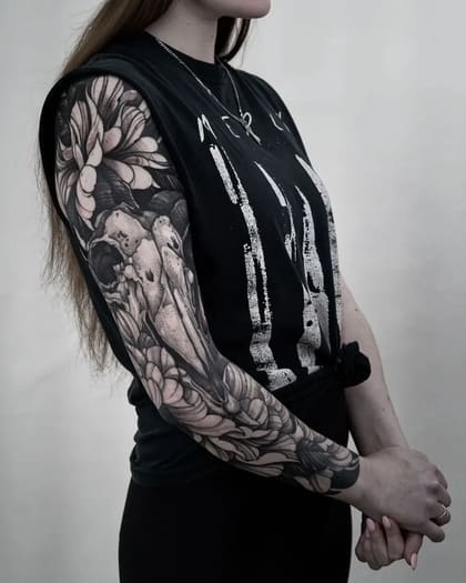 Tatuagem de braço inteiro feminina estilo Blackwork com crânio de animal e flores em tons de preto e cinza.