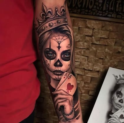 Tatuagem de Calavera Rainha em estilo Chicano e Realismo, com coroa, cartas de ás e rosário, em preto e cinza com olhos azuis e coração vermelho.