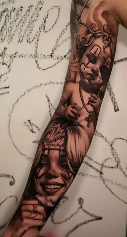 Tatuagem Chicano realista em black and grey de um bebê palhaço com cruz e uma mulher palhaço sorrindo com coroa no braço.