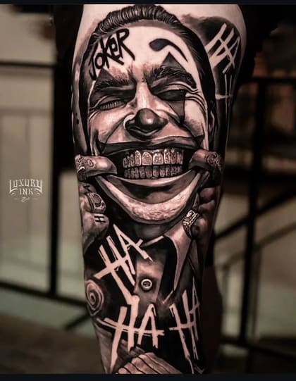 Tatuagem realista do Coringa com maquiagem de palhaço e sorriso macabro, estilo Chicano em preto e cinza na perna.