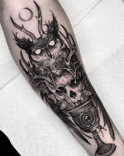Tatuagem blackwork detalhada de coruja com chifres, crânios e cálice místico no antebraço.