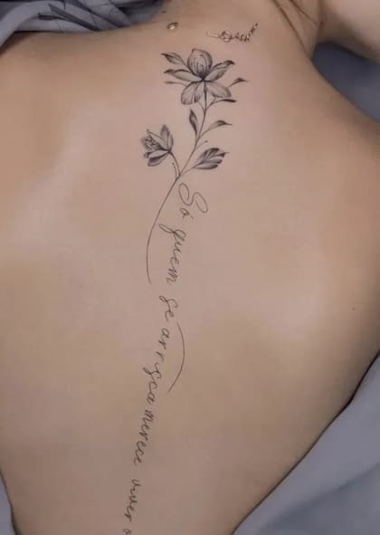 Tatuagem fineline delicada nas costas com flores e a frase 'Só quem se arrisca merece viver o extraordinário'.