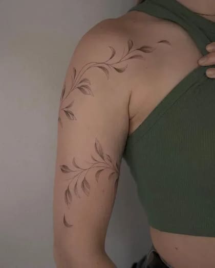 Tatuagem fineline de ramo com folhas delicadas no ombro e braço de uma mulher, em estilo minimalista e orgânico.