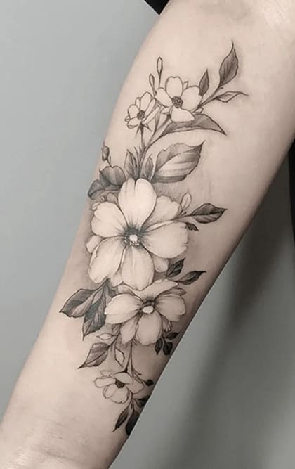 Tatuagem floral delicada em blackwork e fine line no antebraço, com arranjo de flores e folhas em tons de cinza.