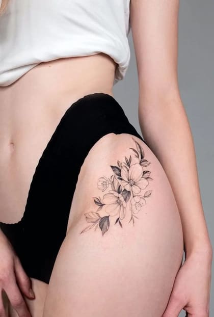 Tatuagem floral delicada em fine line na coxa de uma mulher, com flores e folhas em preto e cinza.