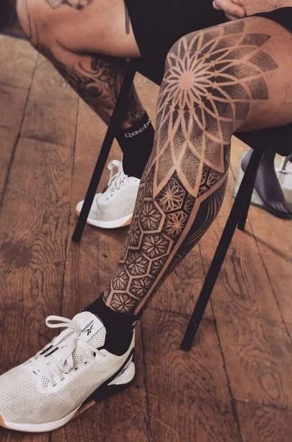 Tatuagem geométrica de pontilhismo na perna, com padrões de mandala e formas hexagonais, em preto e cinza.