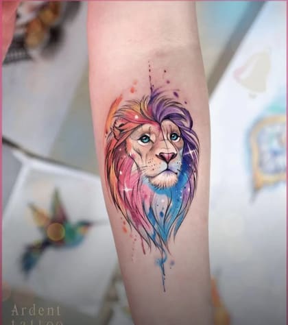 Tatuagem de leão em estilo aquarela colorido no antebraço, com olhos azuis e juba vibrante em tons de rosa, roxo e azul.