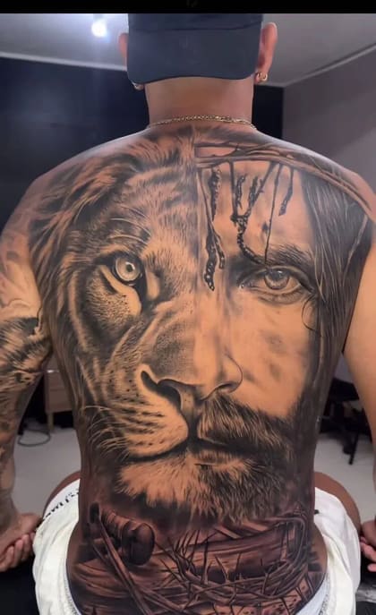Tatuagem de fechamento de costas em realismo preto e cinza, mostrando a fusão do rosto de um leão com o rosto de Jesus Cristo, com coroa de espinhos e pregos na parte inferior.