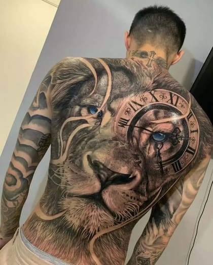 Tatuagem realista de leão com relógio em preto e cinza, olhos azuis, cobrindo as costas de um homem.