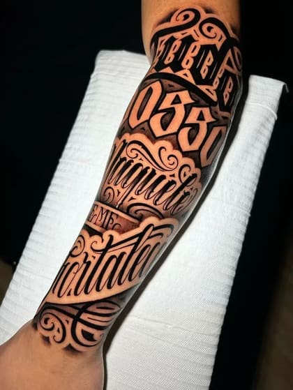 Tatuagem de lettering chicano no braço, com palavras 'Fortaleza' e 'Miguel' em estilo script preto e cinza, com ornamentos.