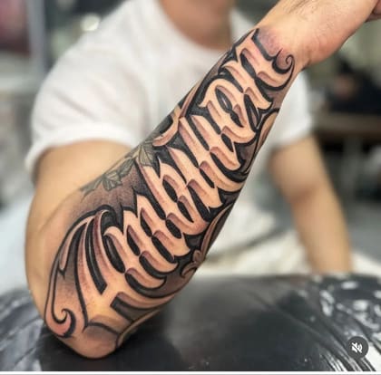 Tatuagem lettering 'Momma' em estilo Chicano 3D no braço, com sombreamento black and grey e detalhes ornamentais.