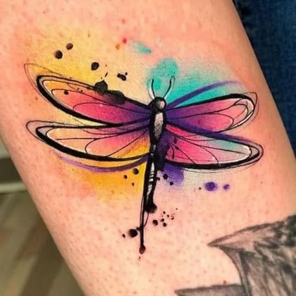 Tatuagem de libélula em estilo aquarela com cores vibrantes como rosa, roxo, amarelo e azul, com contornos pretos, aplicada na pele.