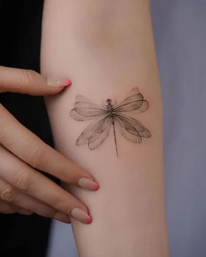 Tatuagem de libélula em estilo fineline no antebraço, com asas translúcidas e detalhes delicados em preto e cinza.