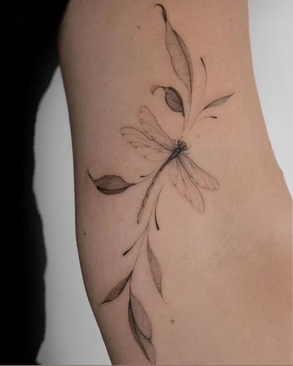 Tatuagem fineline de libélula com asas detalhadas e folhas em estilo dotwork no braço, arte delicada e minimalista.
