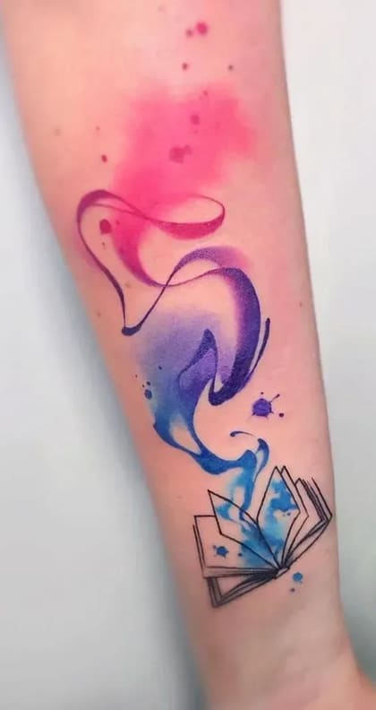 Tatuagem aquarela de livro aberto com tinta rosa, roxa e azul fluindo, no antebraço.