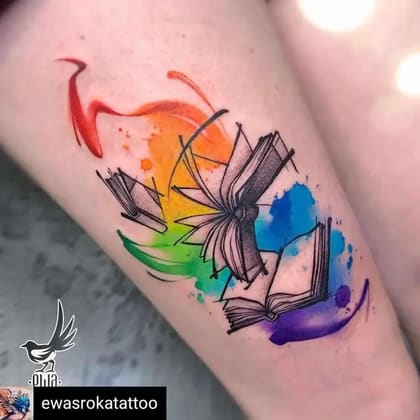 Tatuagem aquarela de livros abertos flutuando com respingos de tinta coloridos em tons de arco-íris, simbolizando conhecimento e imaginação.