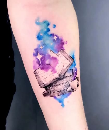 Tatuagem de livros abertos com fundo de aquarela galáctica em tons de azul e roxo no braço.