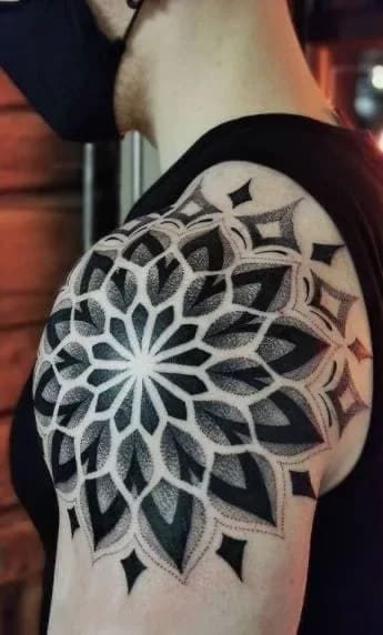 Tatuagem geométrica de mandala em pontilhismo no ombro de um homem, com padrões intrincados em preto e cinza.