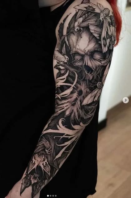 Tatuagem blackwork de manga completa com crânio, mariposas e folhagens em estilo sombrio no braço de uma pessoa.
