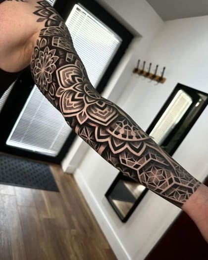 Tatuagem de manga geométrica e dotwork com padrões de mandala em preto e cinza, cobrindo o braço inteiro.