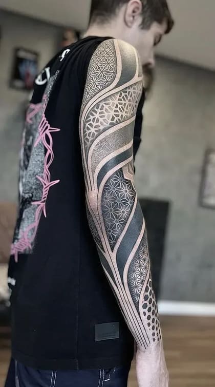 Tatuagem de manga completa no braço de um homem, com design geométrico complexo, padrões de pontilhismo e áreas de blackwork sólido, em tons de preto e cinza.