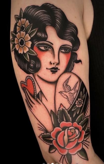 Tatuagem Old School colorida de mulher clássica com flores no cabelo, segurando um coração, com uma andorinha e uma rosa no ombro, em estilo tradicional americano.