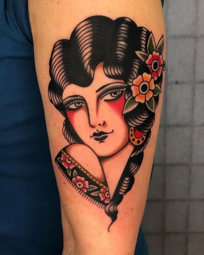 Tatuagem Old School de mulher com flores no cabelo e brinco, estilo tradicional vibrante no braço.