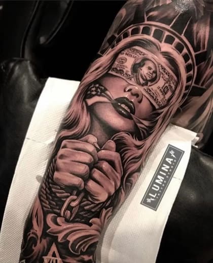 Tatuagem realista de mulher vendada por nota de 100 dólares, com coroa da Estátua da Liberdade, mãos acorrentadas e bandeira dos EUA na boca, em estilo black and grey.