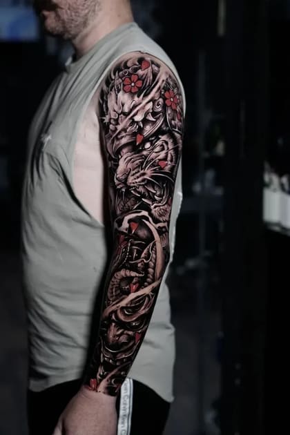Tatuagem de braço inteiro estilo oriental com tigre rugindo, dragão, serpentes e flores de cerejeira em preto e cinza com detalhes vermelhos.