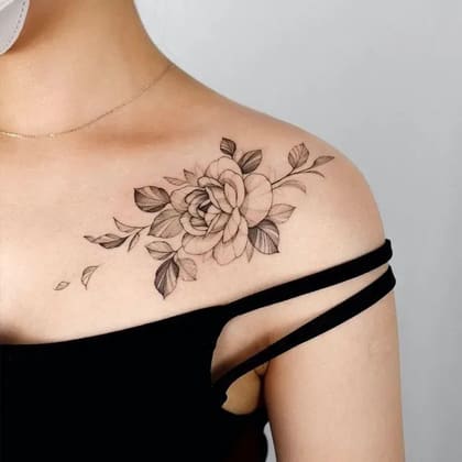Tatuagem floral de rosa em estilo fine line preto e cinza no ombro de uma mulher, com detalhes delicados de folhas e pétalas sombreadas.