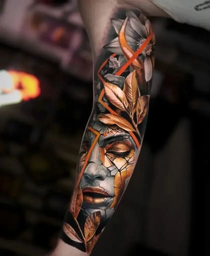 Tatuagem de braço em realismo colorido com rosto feminino de estátua rachada e folhas douradas, estilo surrealista.