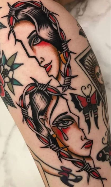 Tatuagem Old School de dois rostos femininos divididos, um sorrindo e outro chorando, envoltos em arame farpado, com cores vibrantes e traços marcantes.