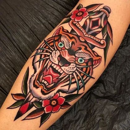 Tatuagem Old School de tigre rugindo com adaga e flores coloridas no braço, estilo tradicional vibrante.