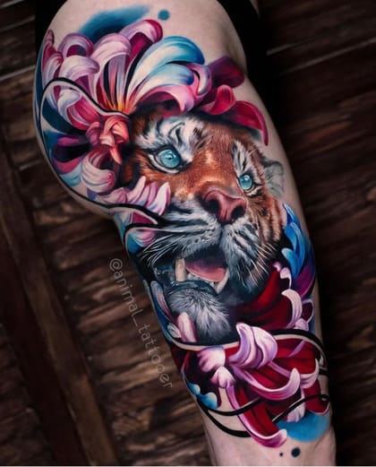 Tatuagem de tigre realista colorido com olhos azuis e flores vibrantes na coxa.