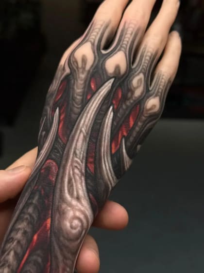 Tatuagem bio-orgânica detalhada na mão e antebraço, com texturas que simulam músculos e ossos em tons de cinza, preto e vermelho.